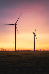 Two wind turbines stand against a vibrant sky during the golden hour, illuminated by warm hues. The colorful sunset creates a stunning contrast with the silhouettes of the turbines.