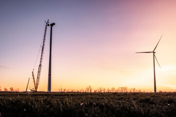 A wind turbine under construction stands beside a completed one during the golden hour. The colorful sky creates a stunning backdrop, highlighting renewable energy in progress.