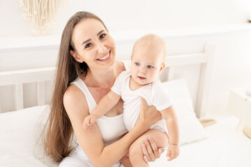 a young beautiful mother with a baby hugs and kisses him or plays with him, having fun on the bed in a bright bedroom, happy motherhood with maternity leave, maternal love and care