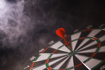 Red Dart Hitting Bullseye on Dartboard Amidst Smoke in Low Light