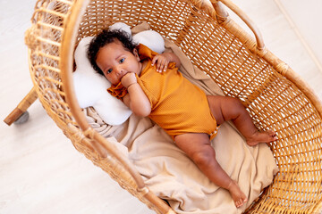 a little African American baby girl in an orange bodysuit in a wicker crib at home has woken up or is falling asleep, a funny six-month-old black newborn baby close-up