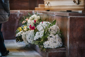 A close-up of floral arrangements placed near a wooden coffin in a funeral setting, symbolizing...