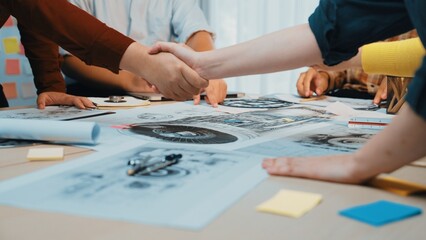 Fototapeta premium A close-up view of hands shaking in agreement over design sketches on a table. The scene depicts teamwork and collaboration in a vibrant workspace filled with creative materials. SACTR