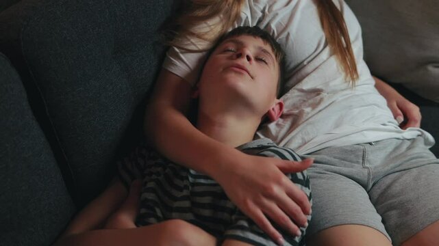 Brother and sister are relaxing on sofa, boy sleeping on her lap, sister gently stroking his hair, creating a peaceful and loving atmosphere