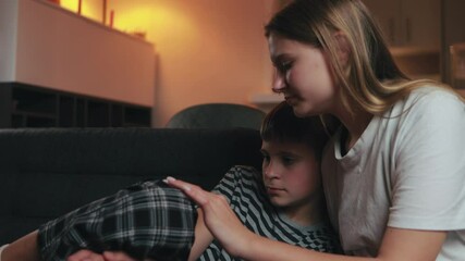 Caring teenage sister gently consoling sad younger brother while sitting together on living room sofa, providing emotional support through compassionate listening and hand stroking