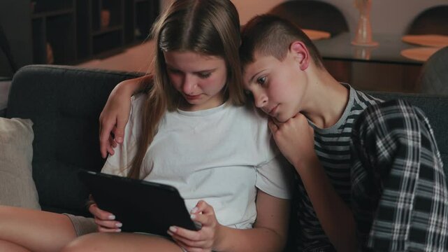 Teenage siblings relaxing together, sharing digital tablet while sitting on comfortable home sofa, engaging in playful interaction and enjoying close familial connection