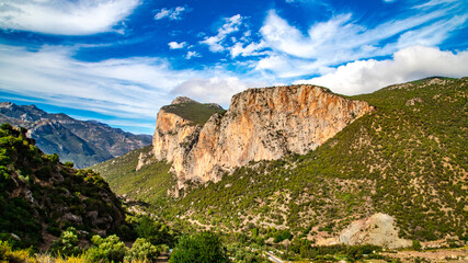 A breathtaking mountainous landscape in Chefchaouen, Morocco, featuring towering rocky cliffs, lush vegetation, and a vibrant blue sky adorned with clouds.