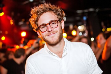 Fototapeta premium Young stylish man with curly hair glasses and beard smiling at vibrant nightlife party scene illuminated by colorful neon lights