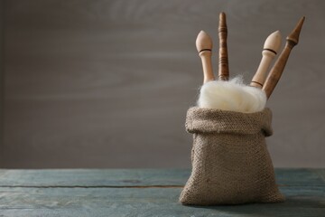 Soft white wool and spindles in burlap sack on blue wooden table. Space for text