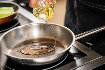 A hand pouring cooking oil into a shiny stainless steel frying pan on a modern stovetop. The scene highlights preparation for cooking in a clean, organized kitchen environment.