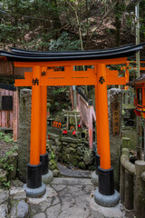 A orange-red torii gate stands at the entrance to a small Shinto shrine nestled in a forested area...