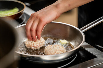 A hand placing breaded patties into a frying pan with butter, showing the cooking process. Another pan with green vegetables is visible, highlighting homemade cuisine.