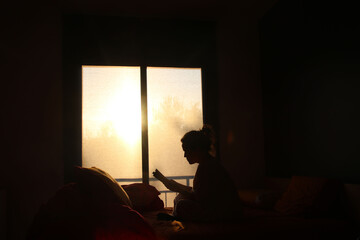 A woman reading a book on her bed in her bedroom. Light streams in through the window at golden hour, and the woman is backlit, her silhouette visible.