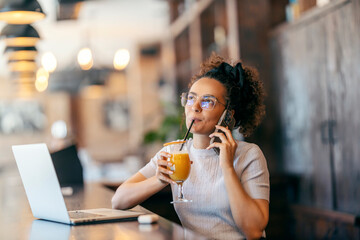A young woman sitting in coffee shop and drinking orange juice while talking on her cellphone.