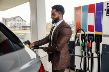 Side view of african american man standing at gas station and filling up car tank.