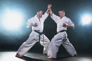 Martial arts masters on dark background. Strong man fighters in white kimono for karate martial arts fighting training in black gym. Karate fighters training punches kicks practicing fight sparring © Юлия Завалишина