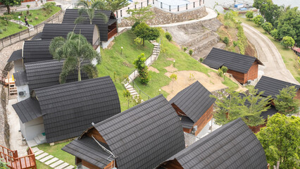 Aerial view of a hillside resort with modern wooden cottages featuring black curved roofs. Green...