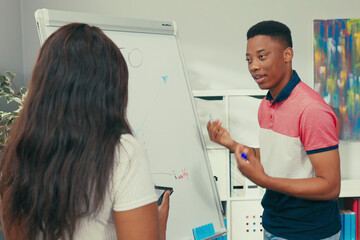Obraz premium Dark-skinned teacher holds tablet in hands stands at blackboard with young handsome student, boy holds marker in hand, looks at graph drawn by translator pointing, school classroom