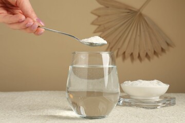 Woman adding collagen powder into glass with water at light textured table against beige...