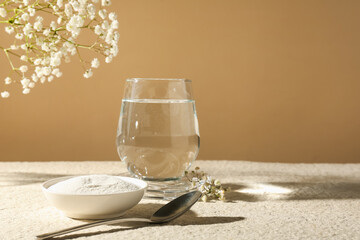 Collagen powder in bowl, spoon, glass of water and gypsophila branches on light textured table against beige background, closeup