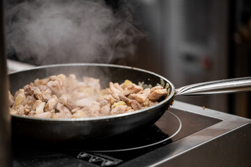 Close-up of a frying pan on a stovetop, cooking pieces of meat. Steam rises, indicating hot food preparation. Captures culinary activity and home cooking atmosphere.