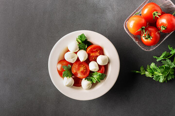 Juicy caprese salad with baby mozzarella cheese, cherry tomatoes and parsley in white ceramic plate on black stone table top view. Healthy diet food concept.