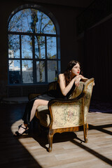Obraz premium An elegant woman in a black dress sits on a sofa. in the background a large window style and beauty Young girl