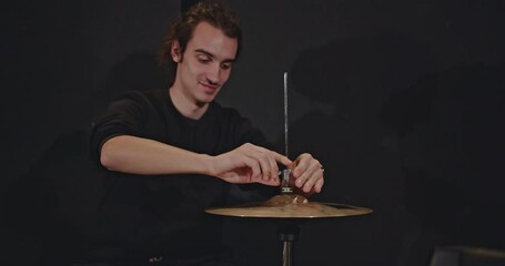 A drummer meticulously adjusts his hi-hat cymbals in a dark studio, preparing for a jazz trio concert. The band practices their music, capturing the essence of live performance.