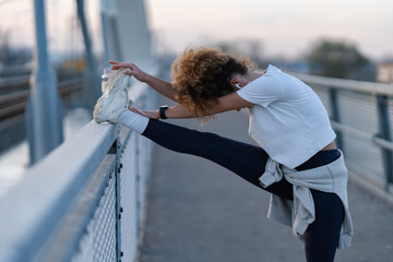 Young woman with curly hair performs a deep leg stretch on a bridge railing during an outdoor...