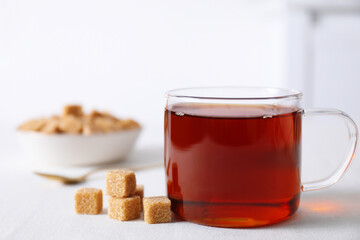 Aromatic black tea in cup and brown sugar cubes on white table, closeup