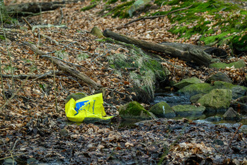A bright yellow bag rests near a flowing stream in a tranquil forest setting. Fallen leaves cover the ground, and moss-covered rocks are visible, reflecting early spring's arrival