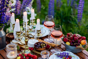 Elegant gorgeous wedding table decor or romantic dinner arrangement outdoors in blooming field. Purple lupine flowers, candles, fruits and wine, wooden vintage furniture. Sunset, summer, golden hour