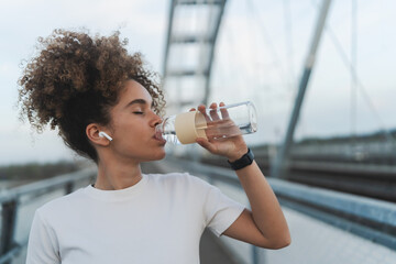 A young woman with curly hair drinks water from a bottle while wearing wireless earbuds and a smartwatch on a bridge. She is dressed in a white t-shirt, staying hydrated after a workout.