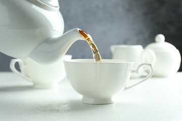 Pouring freshly brewed tea from teapot into cup at white table, closeup