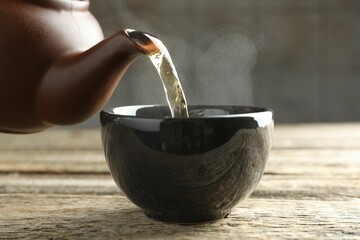 Pouring aromatic tea from teapot into cup at wooden table against grey background, closeup