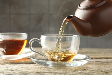 Pouring aromatic tea from teapot into cup at wooden table against grey tiled wall, closeup