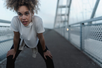 A young woman with curly hair catching her breath after an intense run, leaning forward with hands on her knees, wearing a white T-shirt, leggings, and wireless earbuds on a bridge.