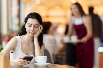 Young woman with mobile phone drinking coffee at table in cafe