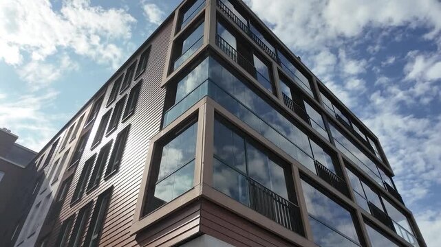 Contemporary urban apartment building with large reflective glass windows and wooden panel siding, captured from a low angle with dynamic sky reflections and soft daylight.