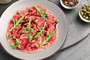 Fresh beef carpaccio with arugula, capers and sauce on dark textured table, above view