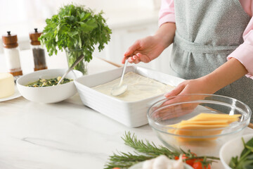 Woman spreading bechamel sauce onto spinach lasagna at marble table indoors, closeup