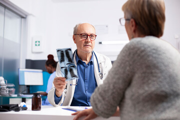 Obraz premium Senior female patient sits across from her retired male physician, who is reviewing xray in hospital office. Elderly doctor discusses medical findings with old woman, outlining treatment options.