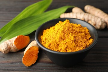 Aromatic turmeric powder in bowl, fresh roots and leaves on wooden table, closeup