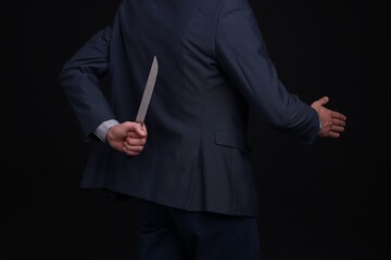 Businessman holding knife behind his back and offering handshake on black background, closeup