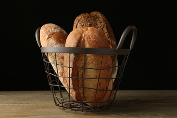 Different types of fresh bread in metal basket on wooden table against black background, closeup
