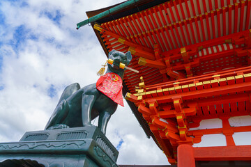 Kyoto, Japan - Sep 23 2024, Close-up view of the stone statue of kitsune, a Japanese fox holding a golden object in its mouth, with red facade of Fushimi-Inari Temple in background, Kyoto, Japan