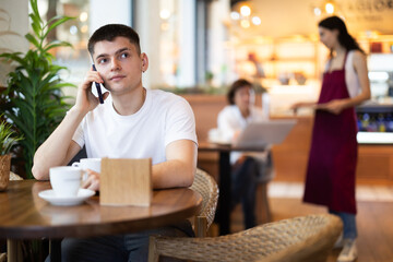 Guy in cafe drinks coffee and using mobile phone, chats online, calls friend, talks on phone. Free Wi-Fi zone, Internet cafe.