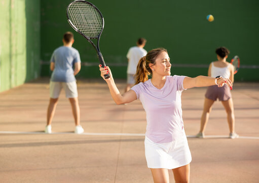 Concentrated young hispanic female frontenis player swinging string racquet to hit ball on outdoor three-walled court on sunny summer day. Sport and active lifestyle concept..