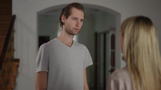 A man in a gray shirt stares silently at his wife, then turns and walks away without saying a word. His serious expression contrasts with her standing, observing his actions. The scene takes place