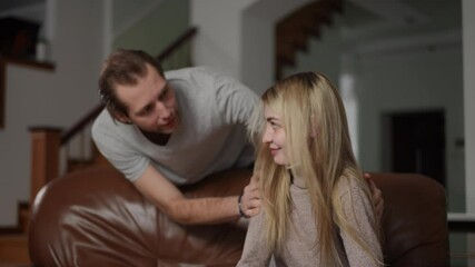 An abusive man standing behind his wife, is angrily speaking while gesturing toward her. She sits on the couch, calmly facing forward, seemingly ignoring him. The scene takes place indoors in a home
